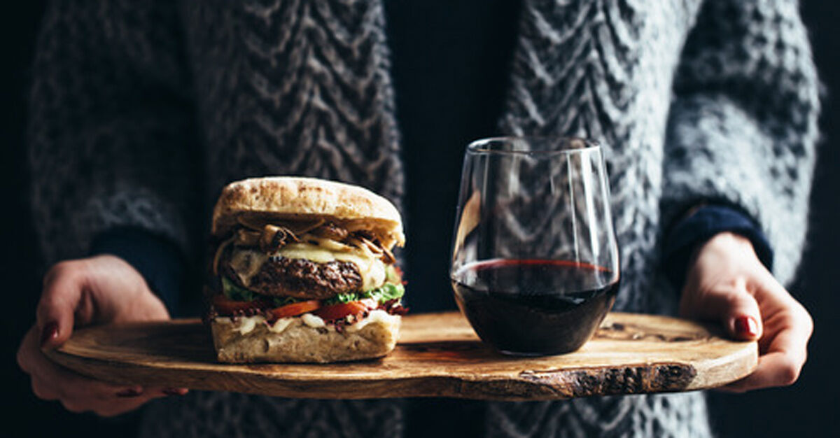 Person holding a wooden platter with a burger and a glass of wine